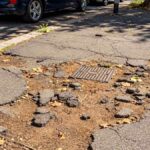 Marciapiede dissestato a Stella Polare, Ostia, con asfalto rotto e chiusino al centro della pavimentazione