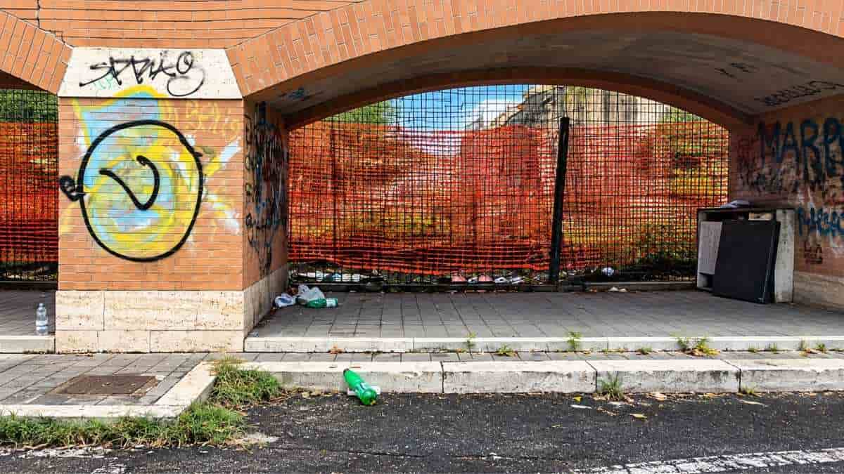 Porticato dell'ex Mira Lanza in via Avogadro, nel quartiere Marconi a Roma, con graffiti, recinzioni arancioni e rifiuti abbandonati sul marciapiede.