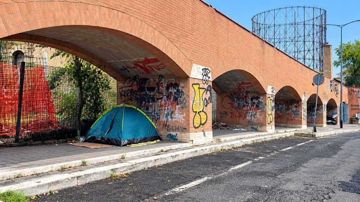 Tenda blu sotto il porticato di via Avogadro, vicino all'ex Mira Lanza nel quartiere Marconi a Roma, tra graffiti e recinzioni di cantiere.
