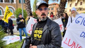 Manifestante con megafono e maglia di Giustizia X Ostia, circondato da persone e striscioni durante una protesta a Ostia.