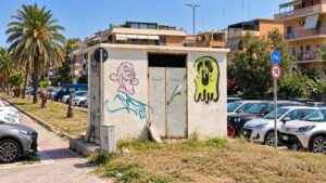 Cabina elettrica di viale delle Repubbliche Marinare a Ostia, con graffiti sulle pareti, porte metalliche e automobili parcheggiate nelle vicinanze.