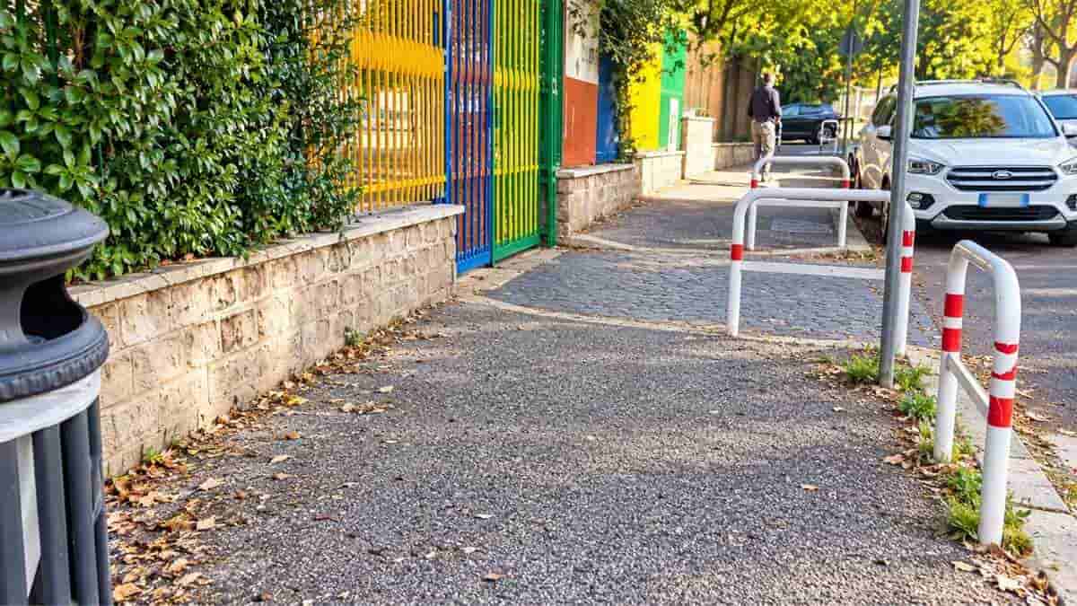 Roma, percorso pedonale con pavimentazione deteriorata e barriere protettive vicino a una scuola nel quartiere Tor Marancia.